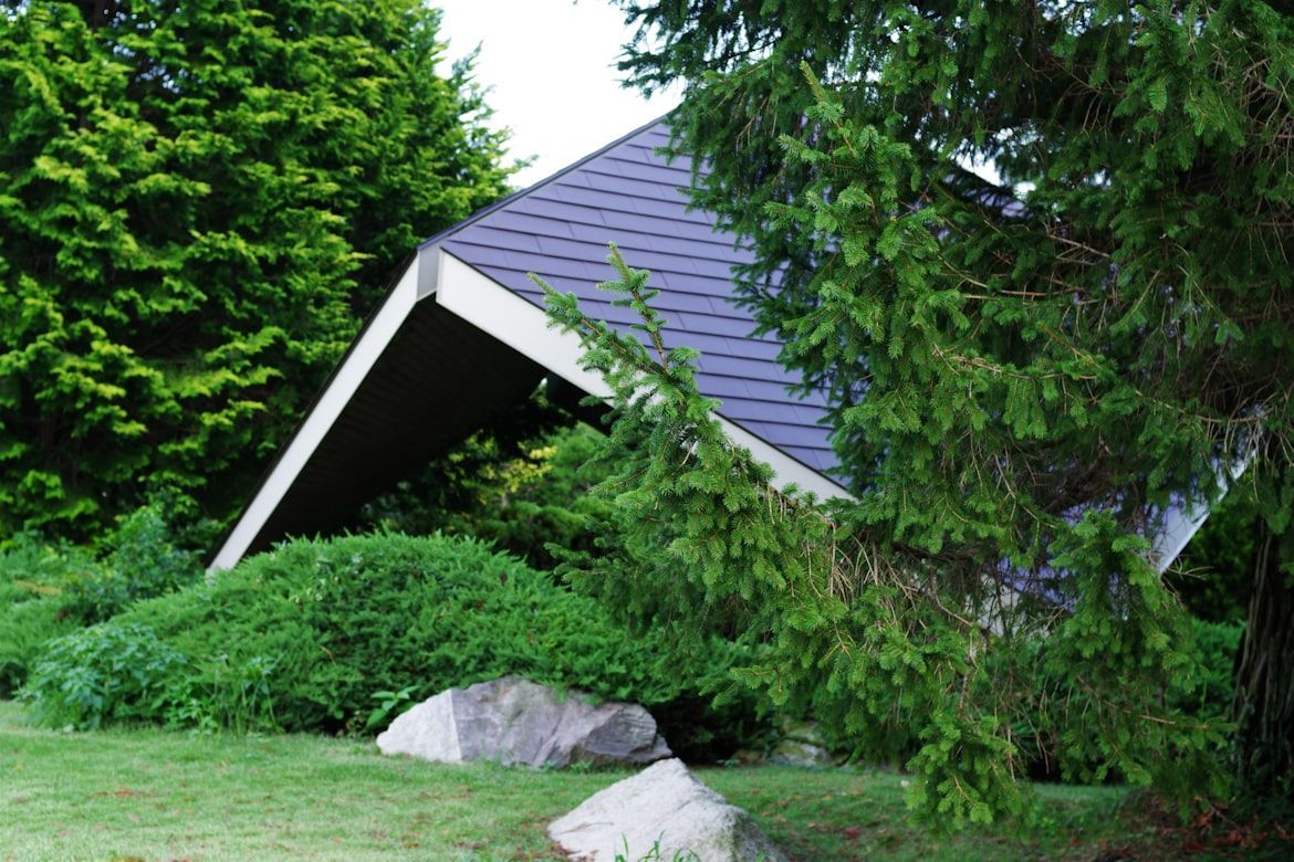 Triangular roof structure with dark shingles amidst green shrubs and trees.