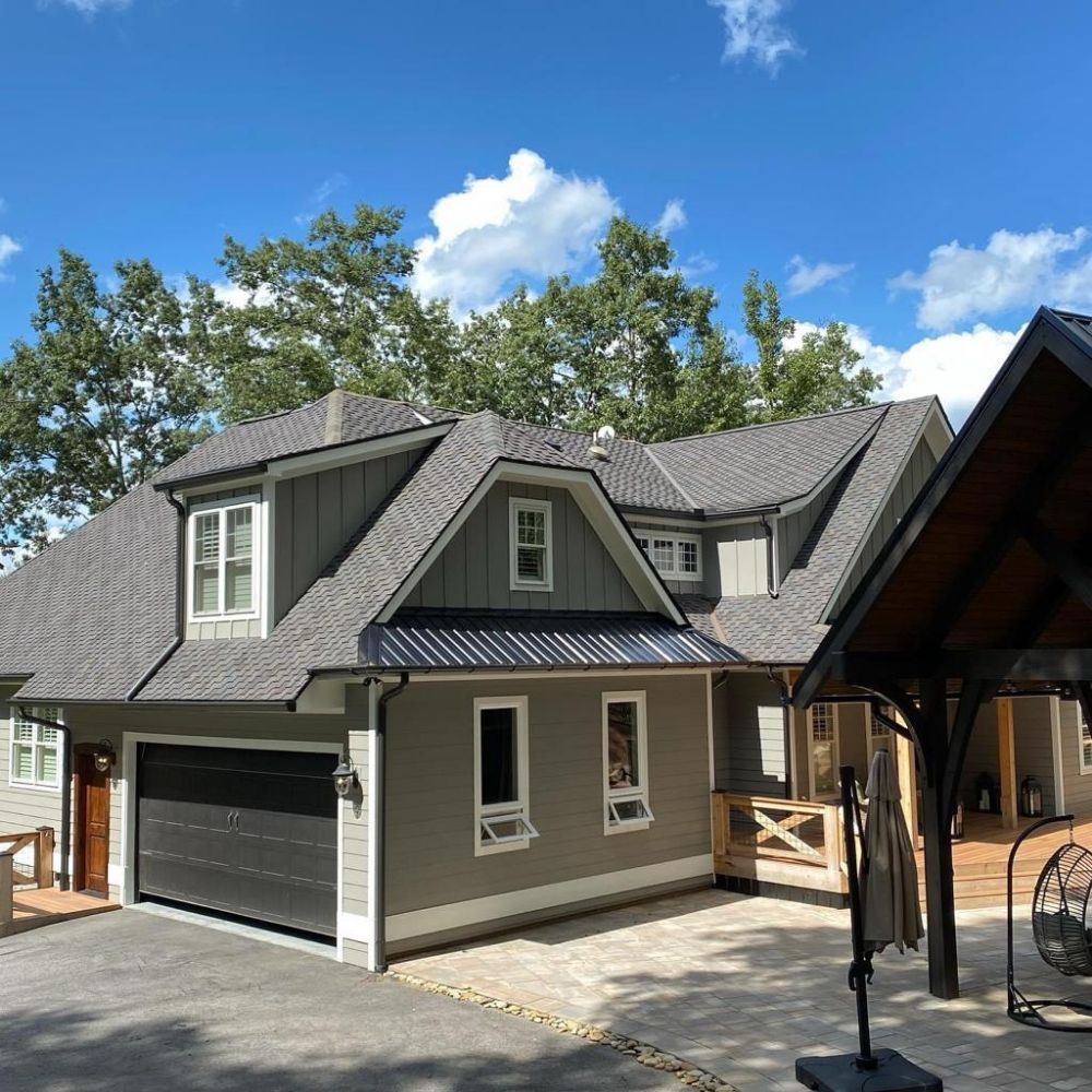 Gray house with dark garage door, and a black awning. Blue sky with trees in the background.