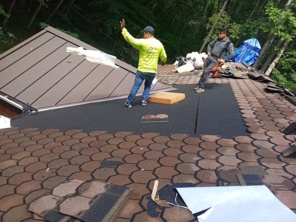 Two roofers working on a roof, one waves. Brown shingles, gray metal, and forest background.