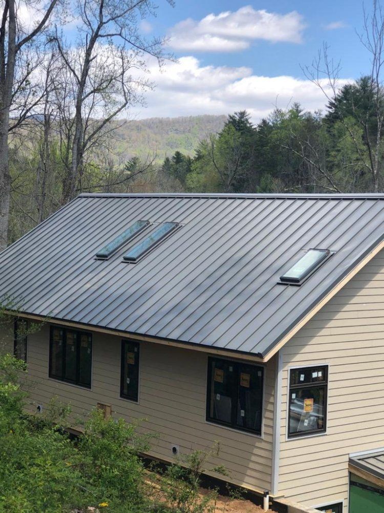 A house with a gray metal roof and three skylights, tan siding, and black window frames, set against a mountainous backdrop.