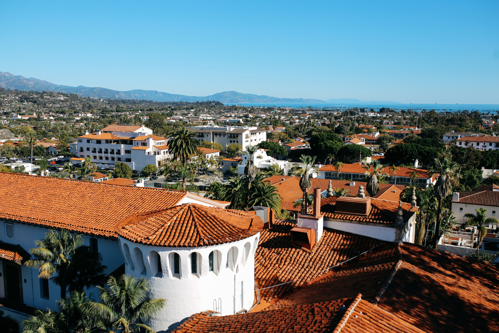 Cityscape with terracotta roofs, white buildings, mountains, and the ocean under a clear blue sky.