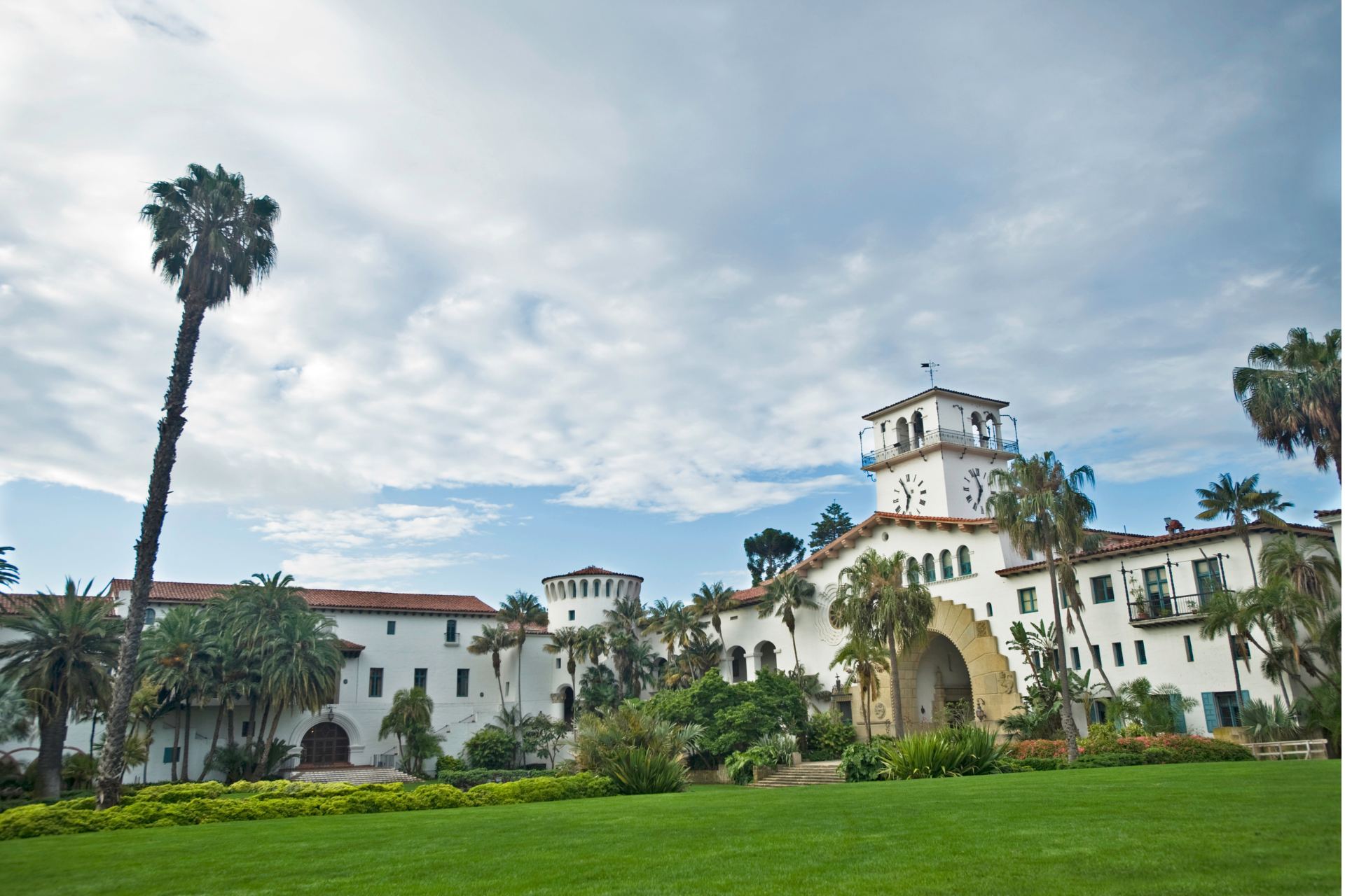 Santa Barbara Courthouse, white stucco building with red tile roof, palm trees, and green lawn under cloudy sky.