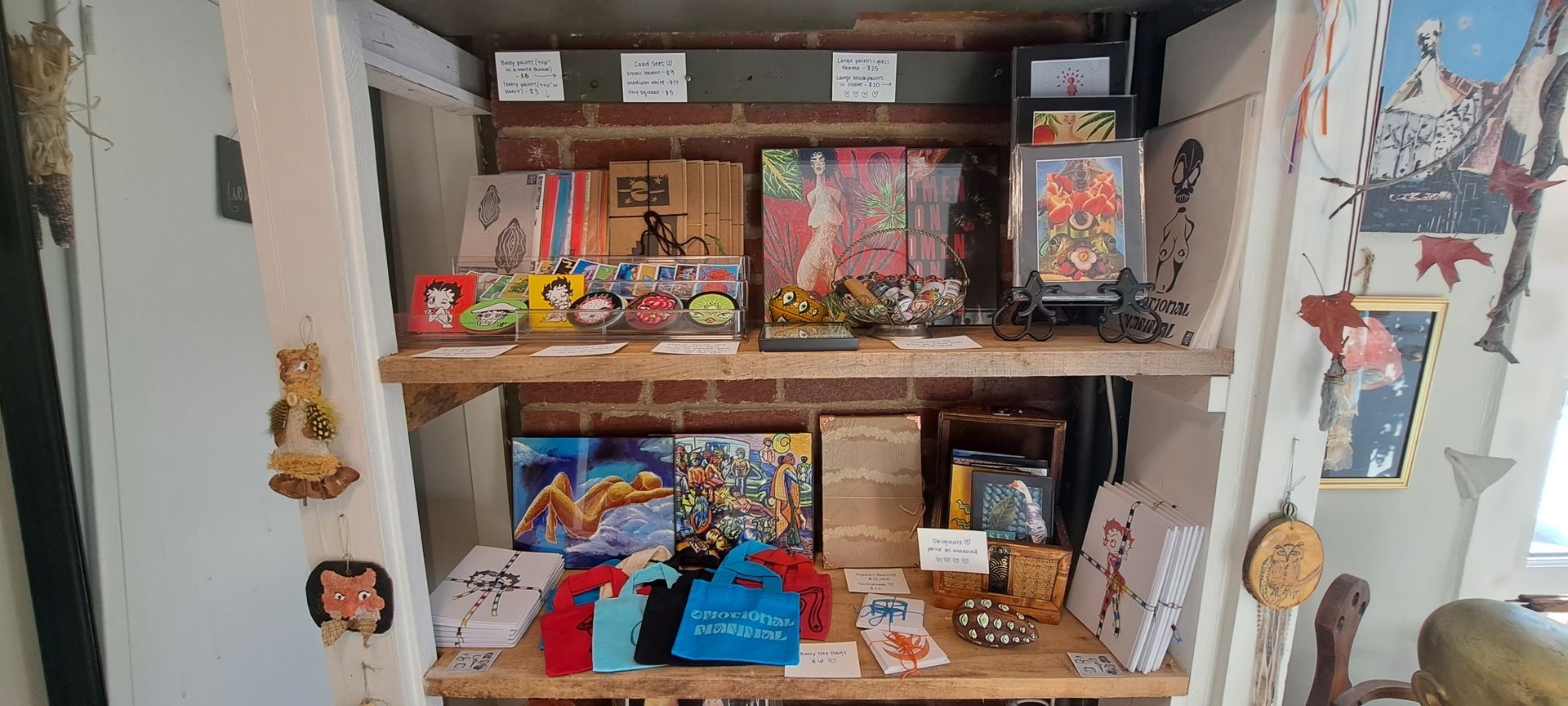Shelves displaying various items for sale in a shop. Artwork, toys, bags, and other merchandise are visible.