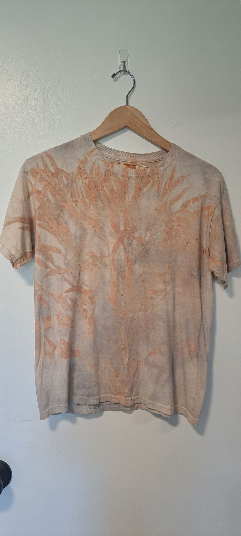 A tie-dye short-sleeved shirt with an orange design hangs on a wooden hanger from a hook on a white wall.