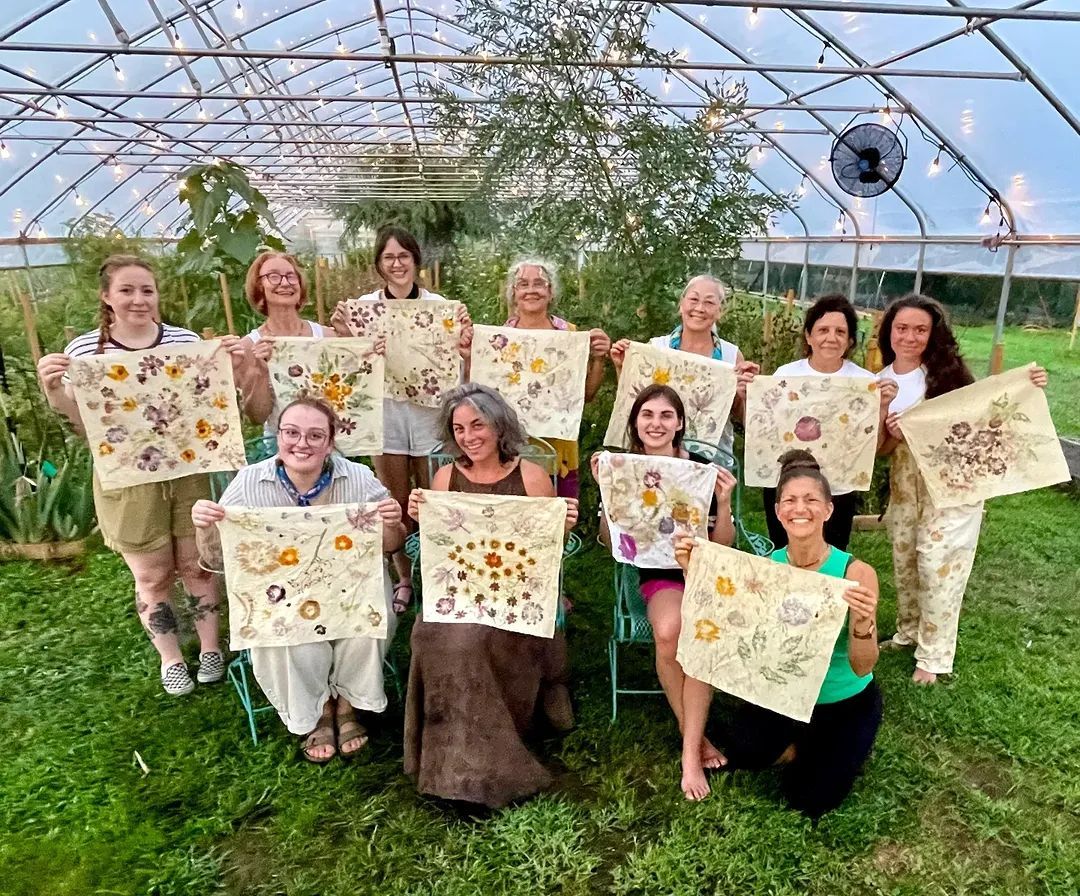 Group of women holding fabric art with pressed flowers, gathered in a greenhouse.
