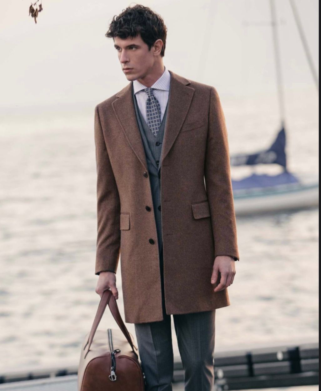 Man in brown overcoat, suit, holding bag, near water with boat.