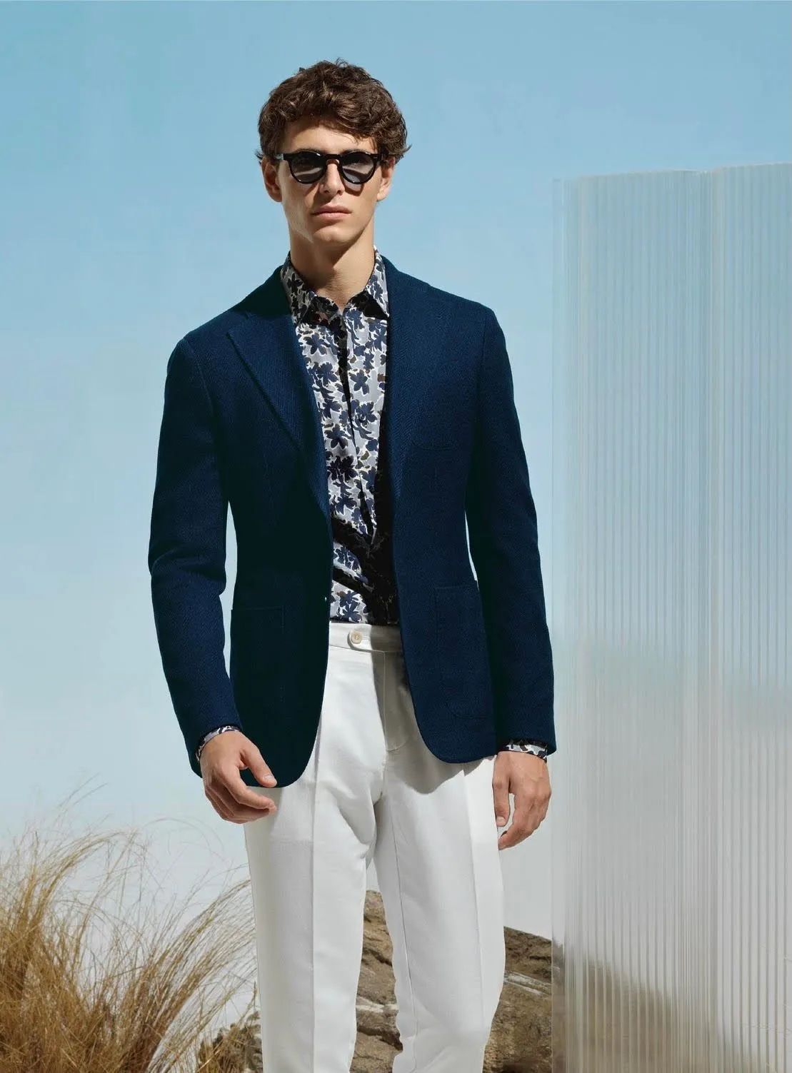 Man in navy blazer, floral shirt, white pants, sunglasses, posing outdoors.