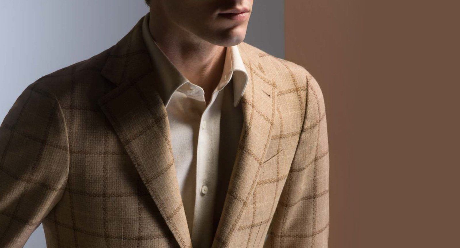 A man wearing a suit jacket and a white shirt is standing in front of a brown wall.