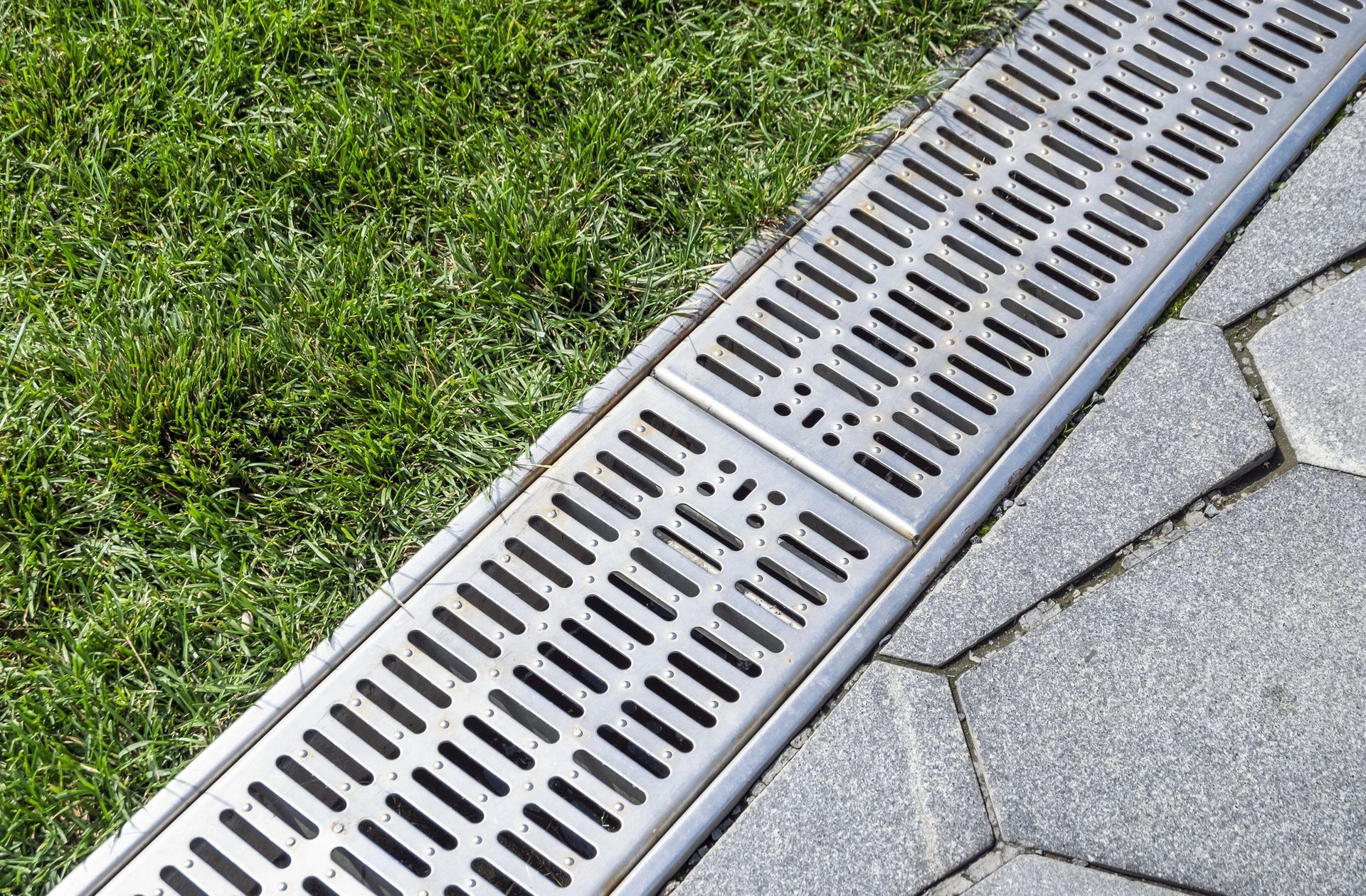 Metal trench drain on a gray stone path next to green grass.