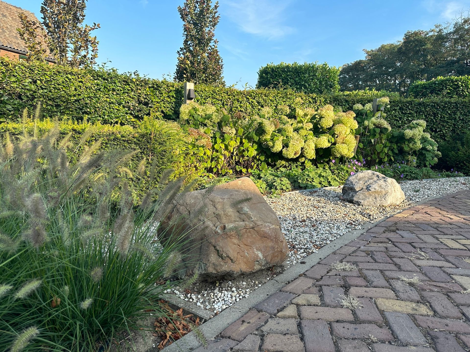 Lush garden with large rocks, ornamental grasses, and manicured hedges. Brick path in foreground, blue sky.