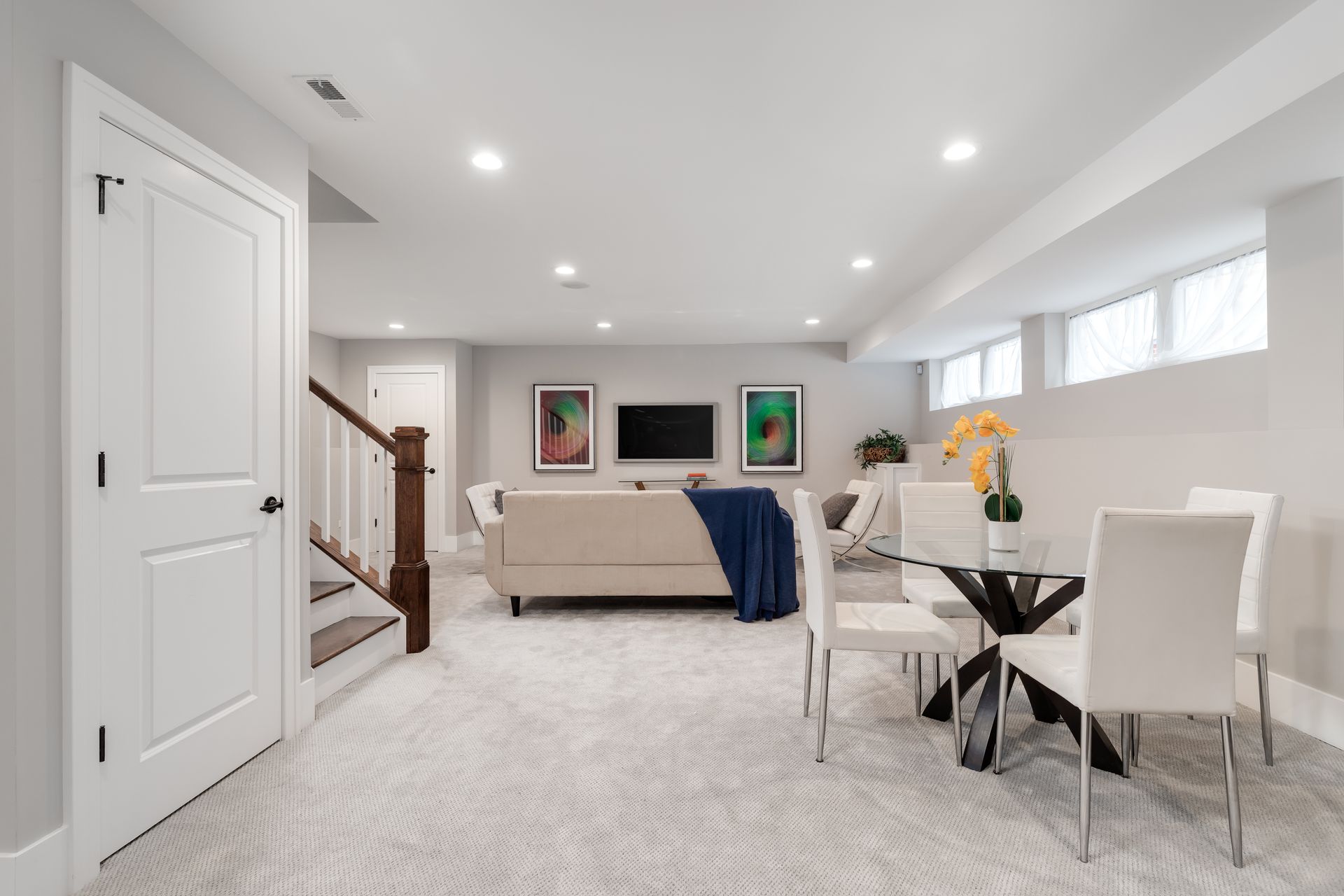 Modern basement with living and dining areas; beige sofa, white chairs, TV, and a staircase.