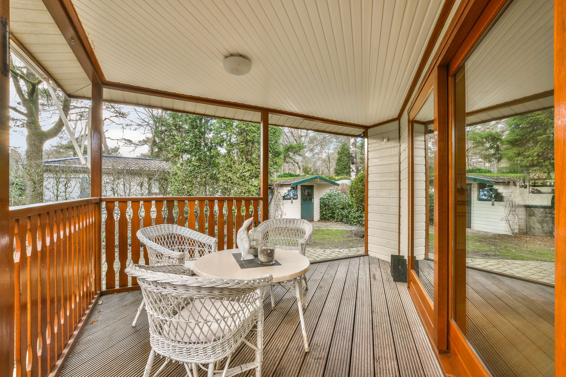 A porch with white wicker furniture. Brown wooden railings, trim, and flooring. Overlooking trees and a small building.