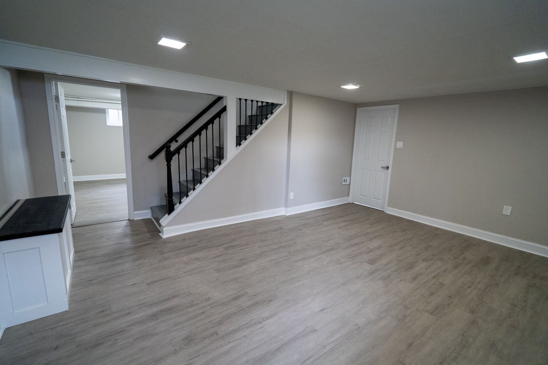 Empty, renovated basement with light wood flooring, stairs, and white walls.