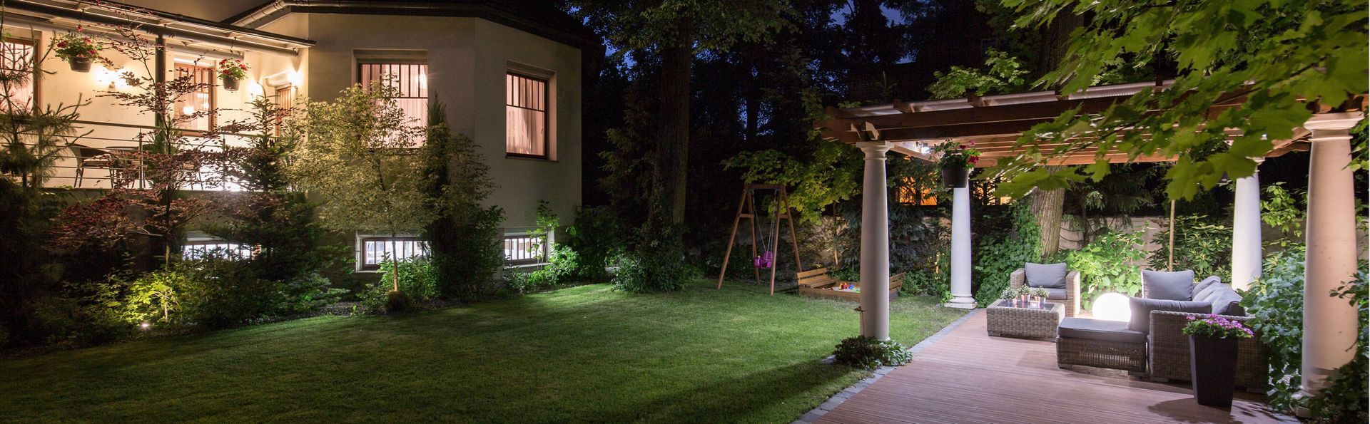 Night view of a backyard with a house, lawn, and a pergola with seating. Warm lights illuminate the scene.