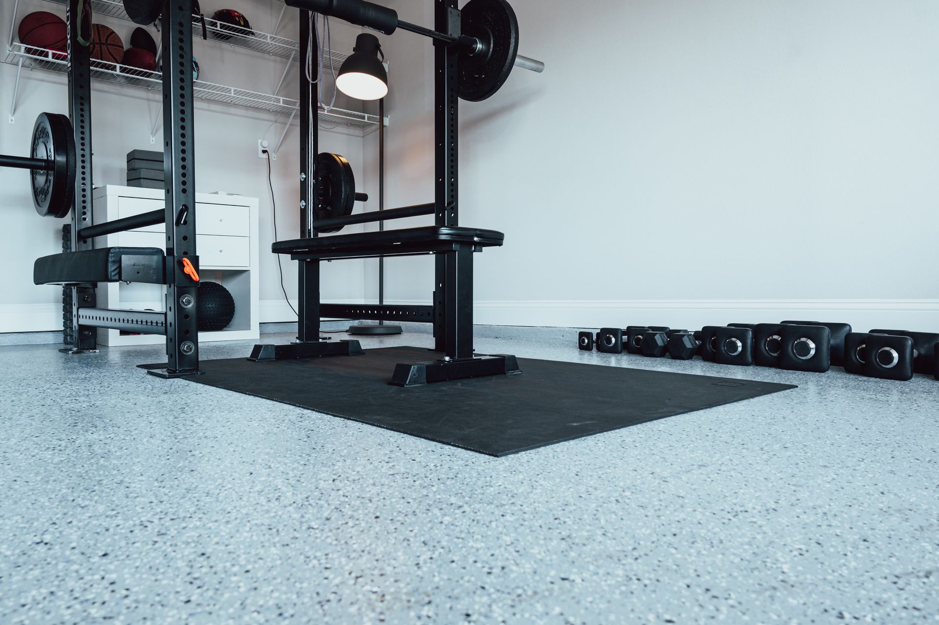 Garage gym with weight rack, weights, and black rubber flooring on speckled concrete.
