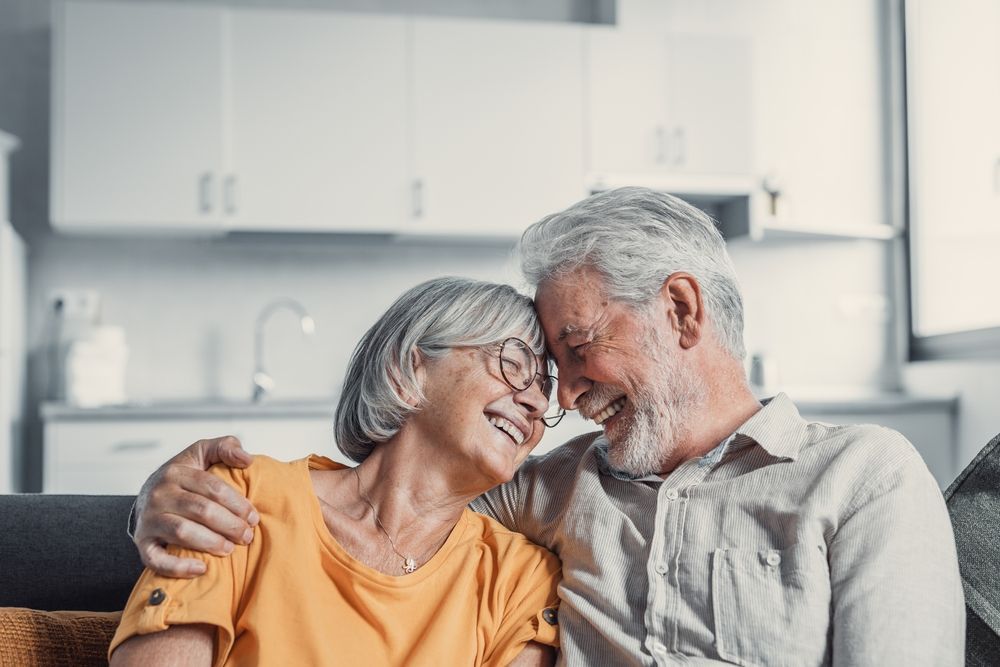 Elderly couple smiling, embracing, and touching foreheads; sitting on a sofa in their home.