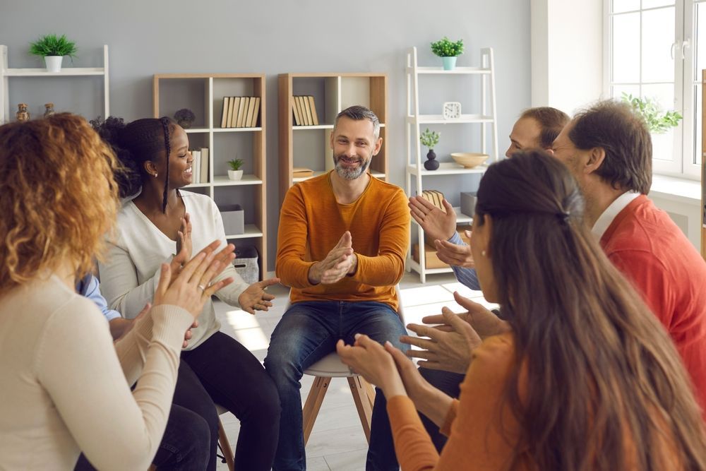 People sitting in a circle, clapping for a smiling man in an orange sweater.