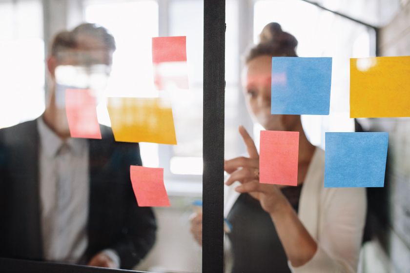 A man and a woman are looking at sticky notes on a glass wall.