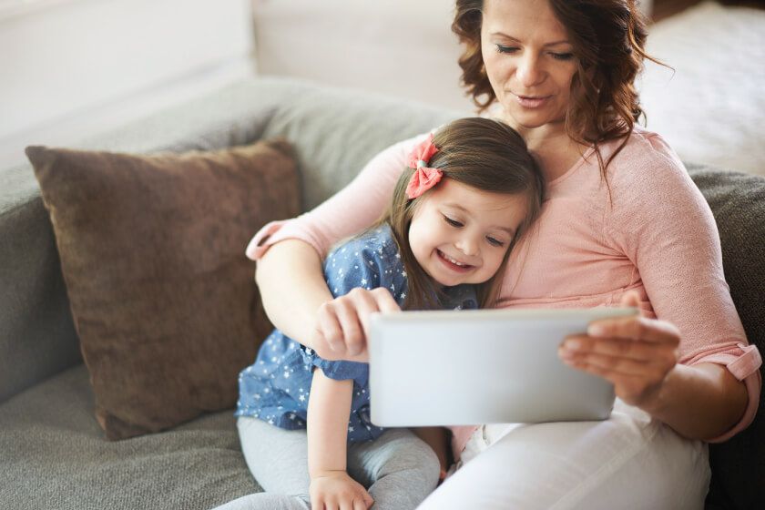 A woman and a little girl are sitting on a couch looking at a tablet.