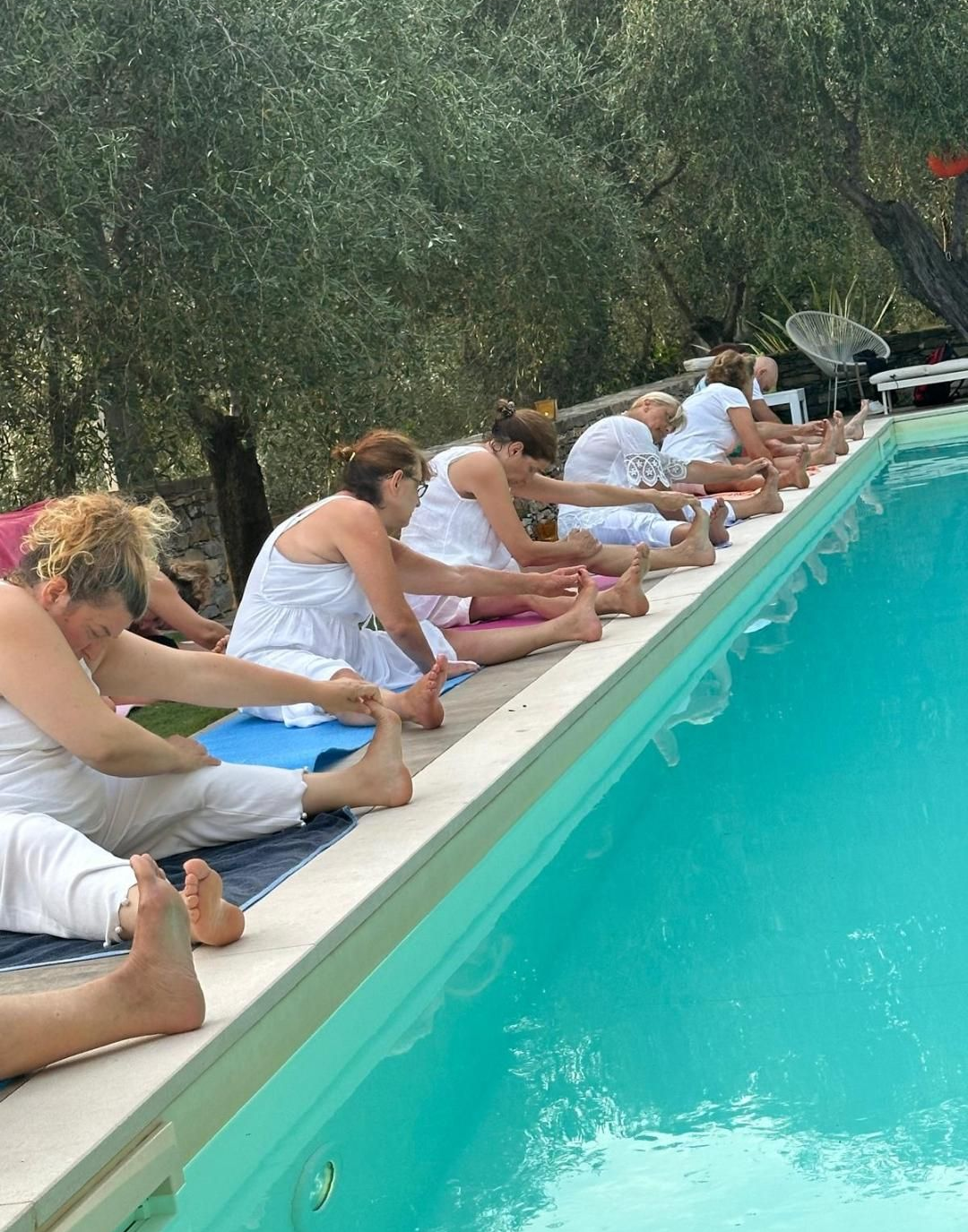 Persone vestite di bianco si stendono vicino a una piscina, sullo sfondo degli ulivi.