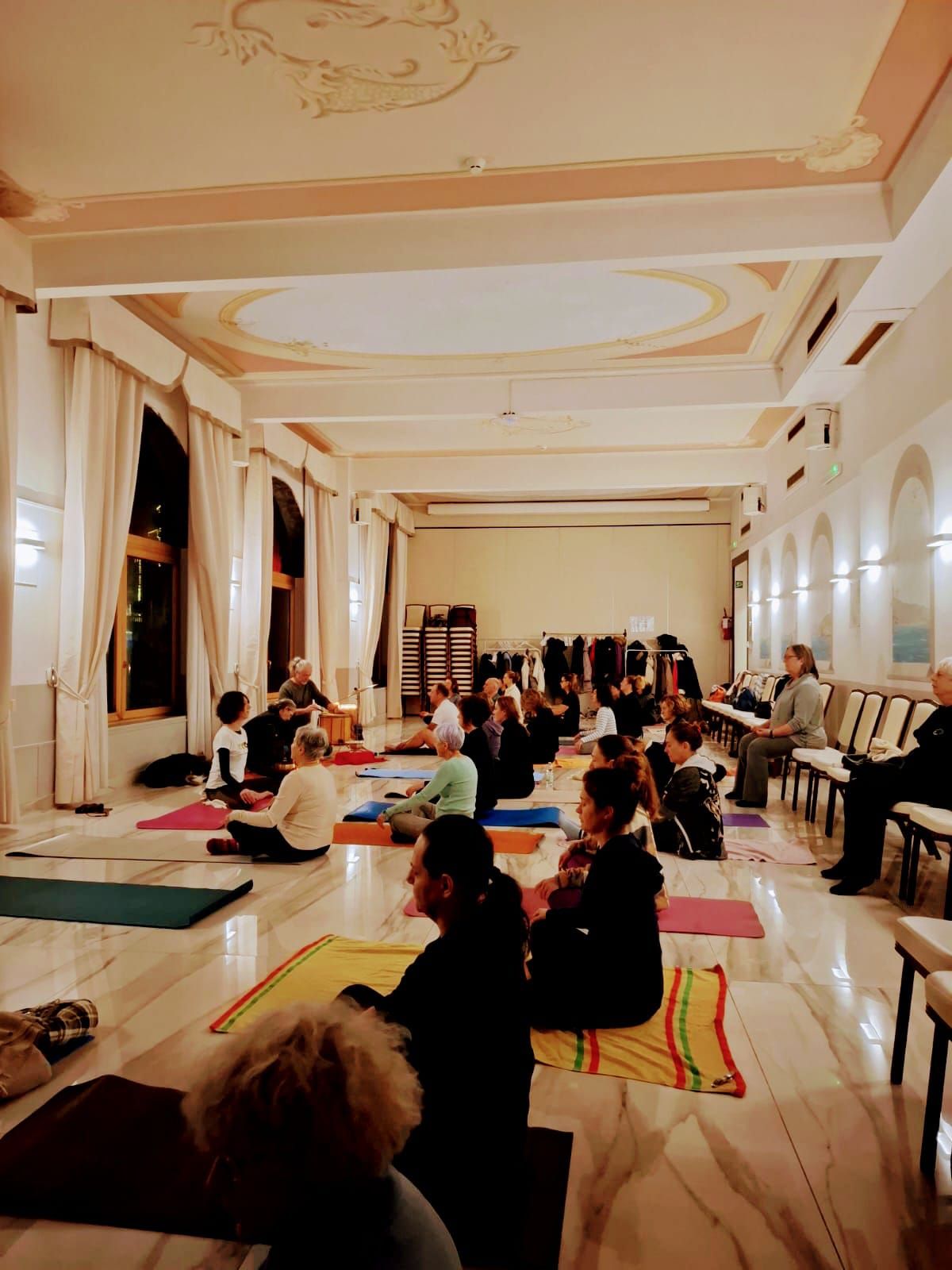 Persone in una grande sala che praticano yoga su tappetini. Soffitto decorato e colonne, con finestre su un lato.