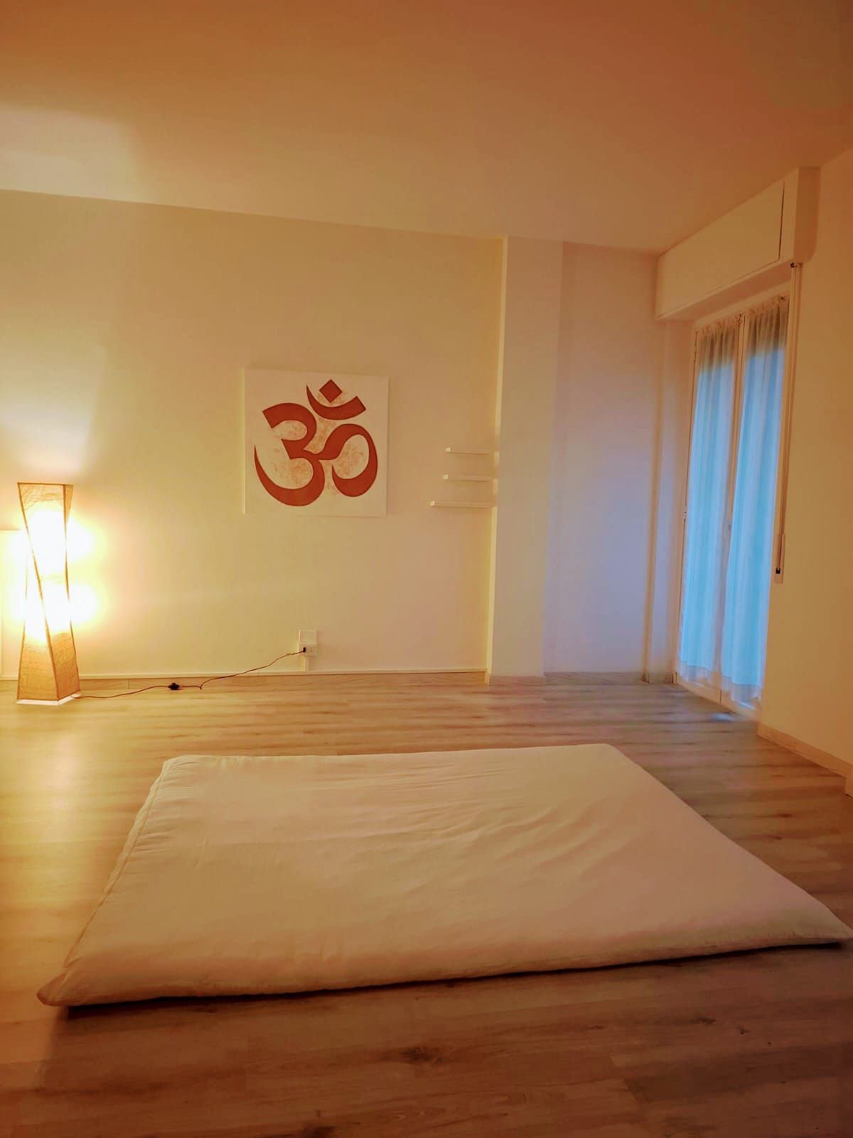 Sala yoga con tappetino, simbolo Om, lampada da terra e finestra con tende.