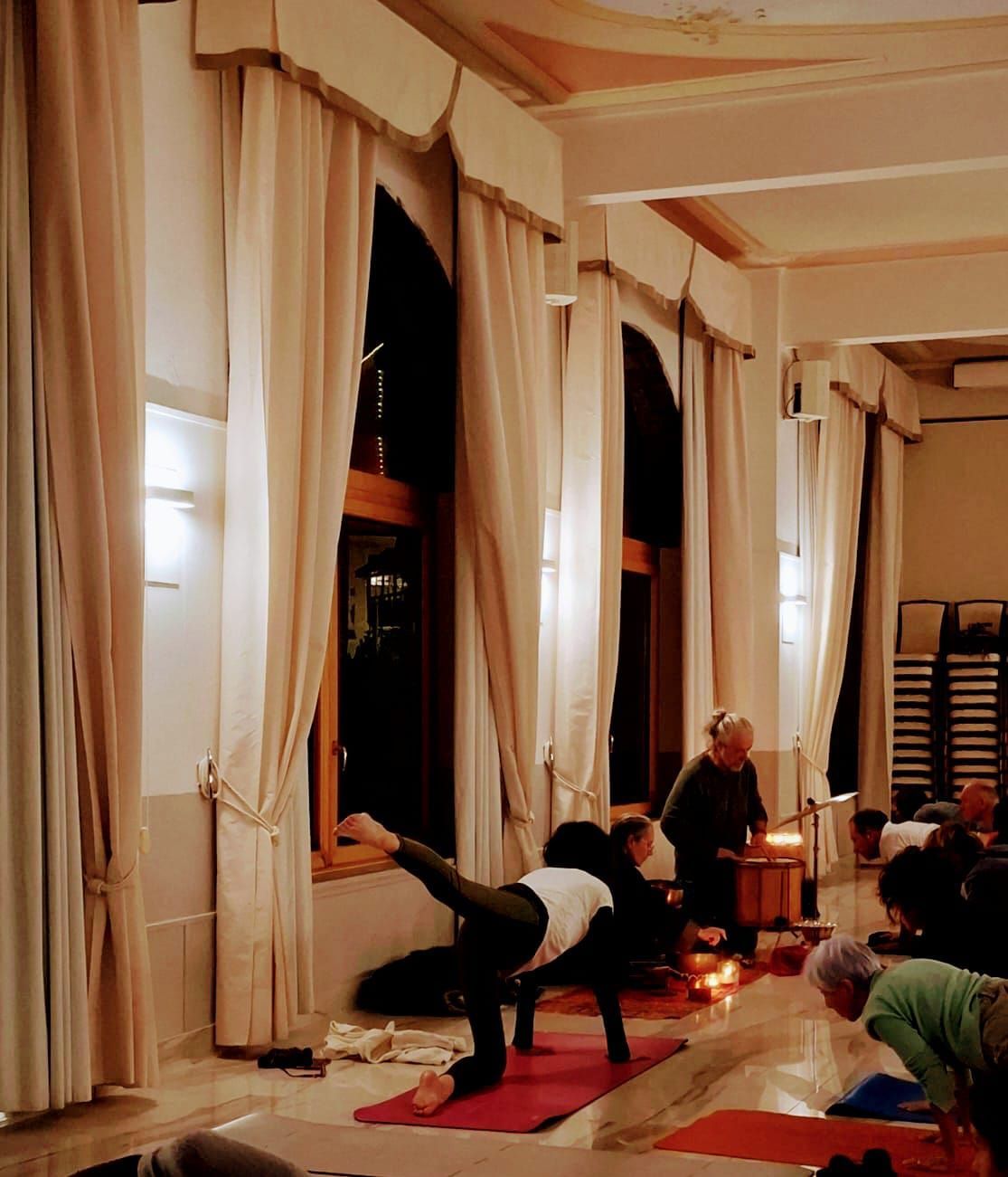 Lezione di yoga in una stanza con alte finestre e tende. Persone sui tappetini, alcune in posizioni. Luci soffuse.