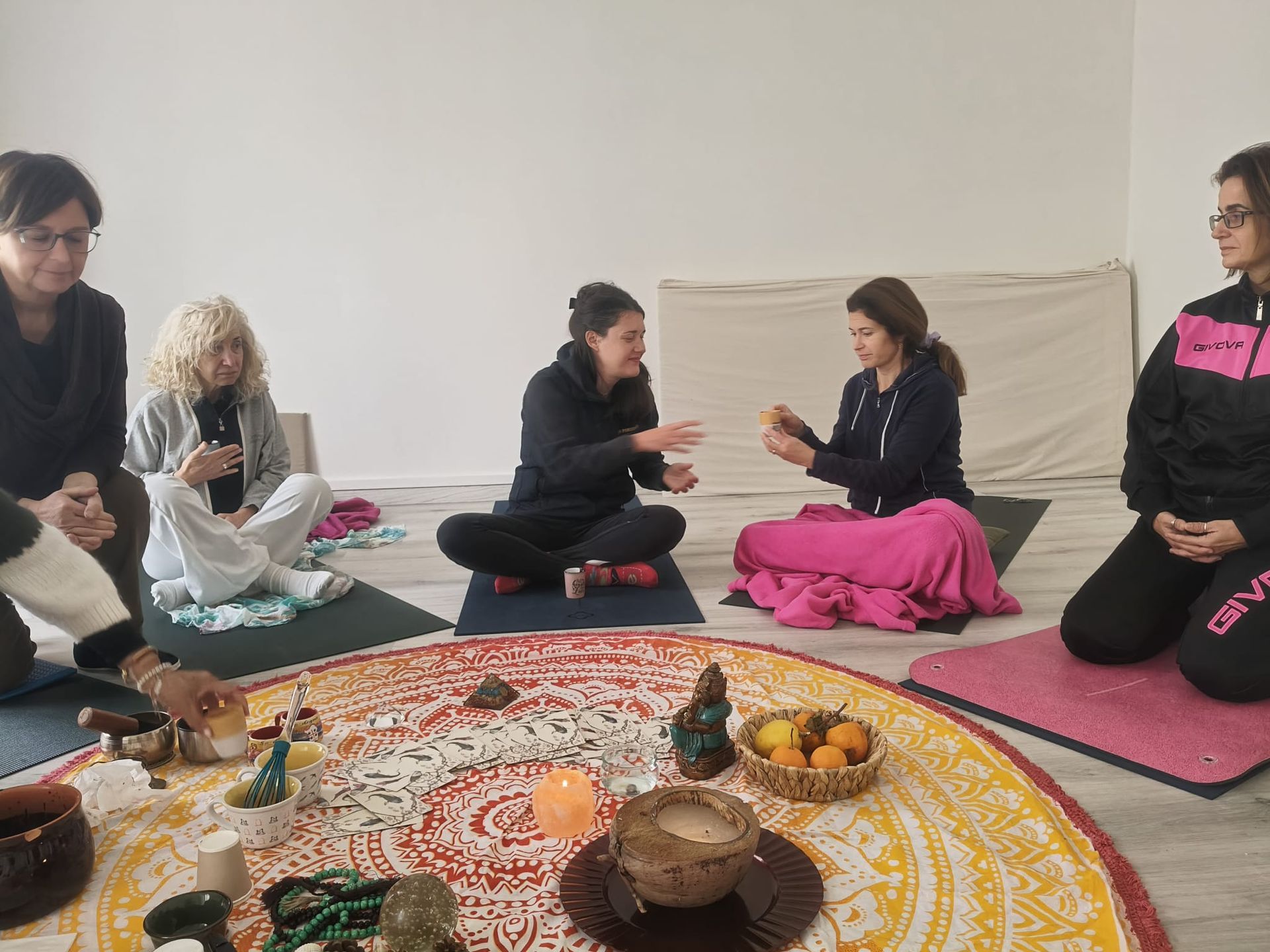 Le persone siedono in cerchio su stuoie, meditando e tenendo in mano oggetti. Al centro ci sono un tappeto mandala e delle candele.