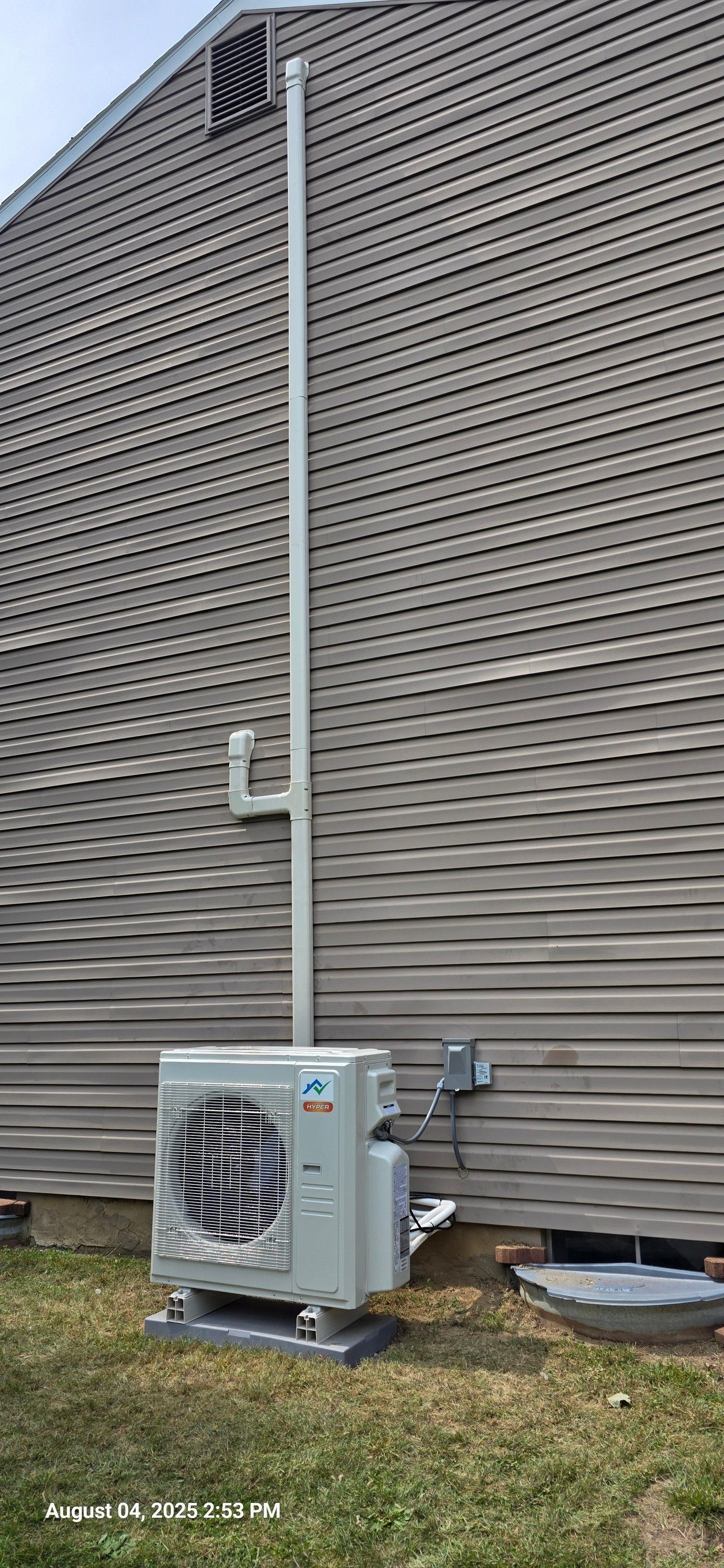 An exterior air conditioning unit attached to a house with a white pipe running up the siding.