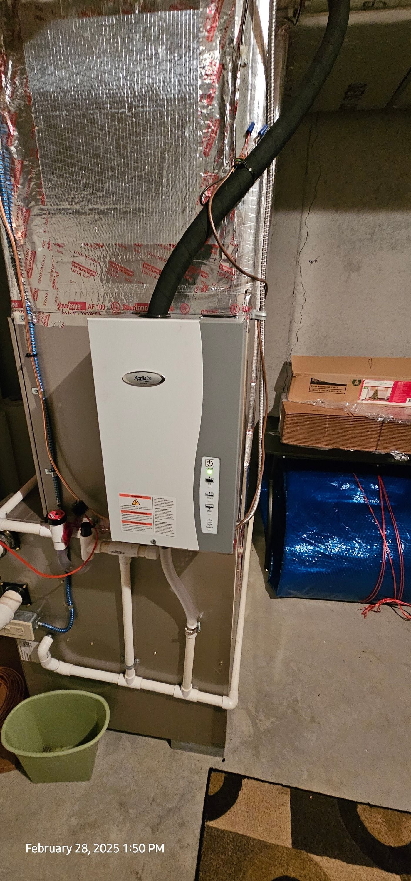 An air handler unit with ductwork in a basement setting. A green bucket sits in the foreground.