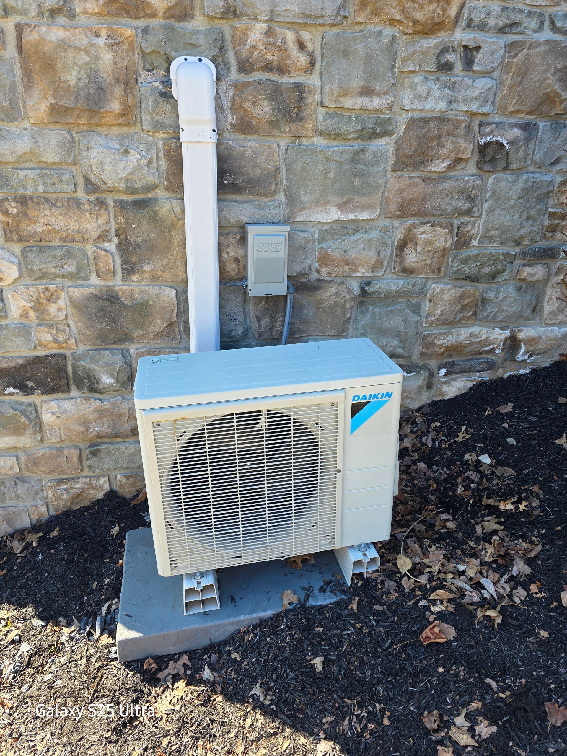 Air conditioning unit mounted on a concrete pad near a stone wall, with white vent pipe.