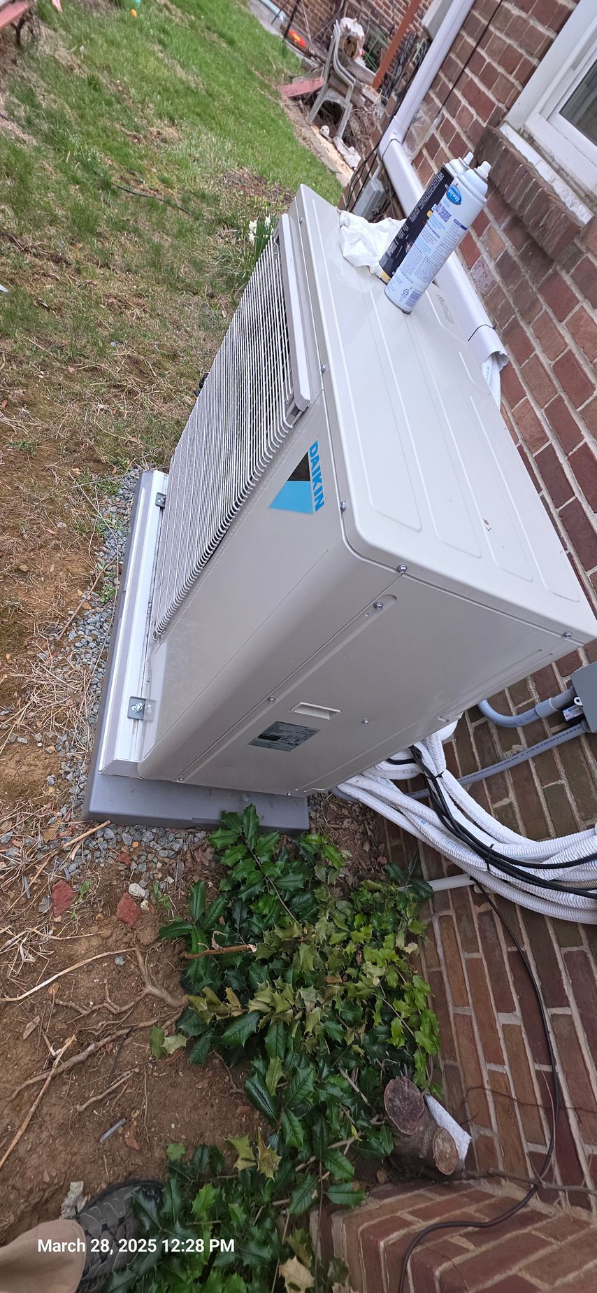 An air conditioning unit mounted on a concrete slab near a brick wall, surrounded by foliage.