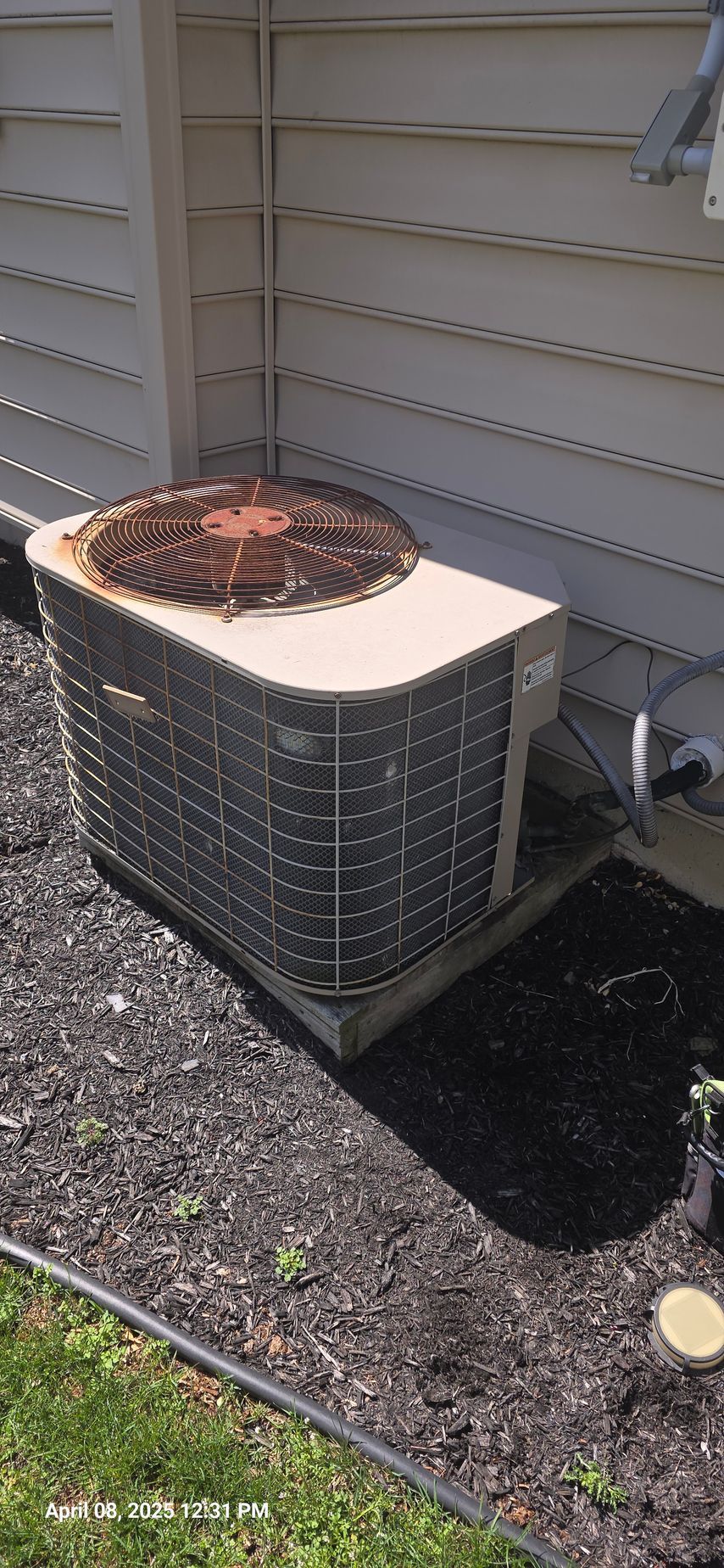 An outdoor air conditioning unit next to a light-colored building with dark mulch and grass in front.