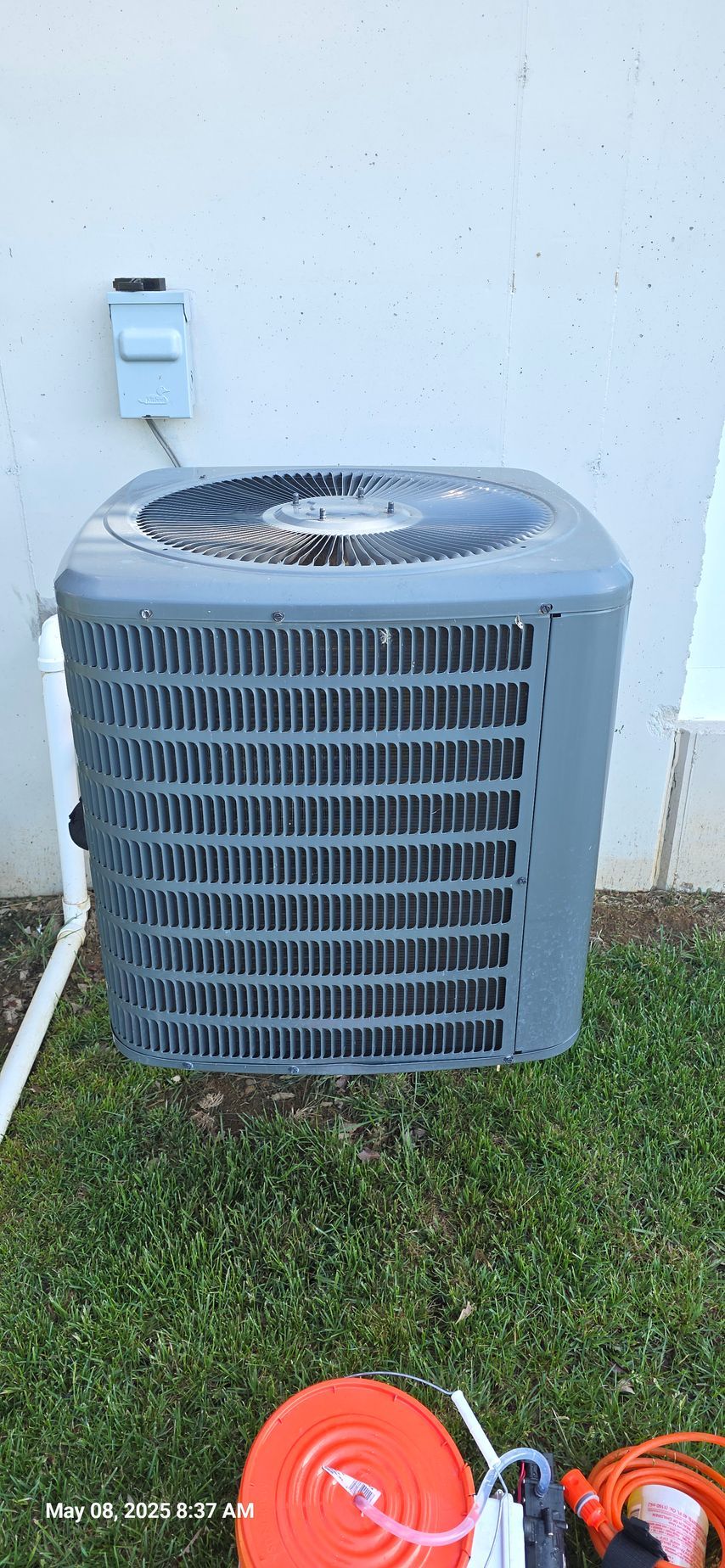 An outdoor air conditioning unit with a gray exterior sits on grass next to a white wall.
