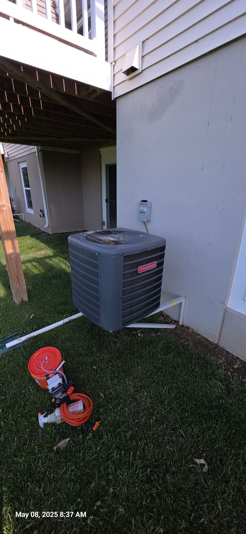 An air conditioning unit next to a house under a deck. Orange reel and tools are on the grass.