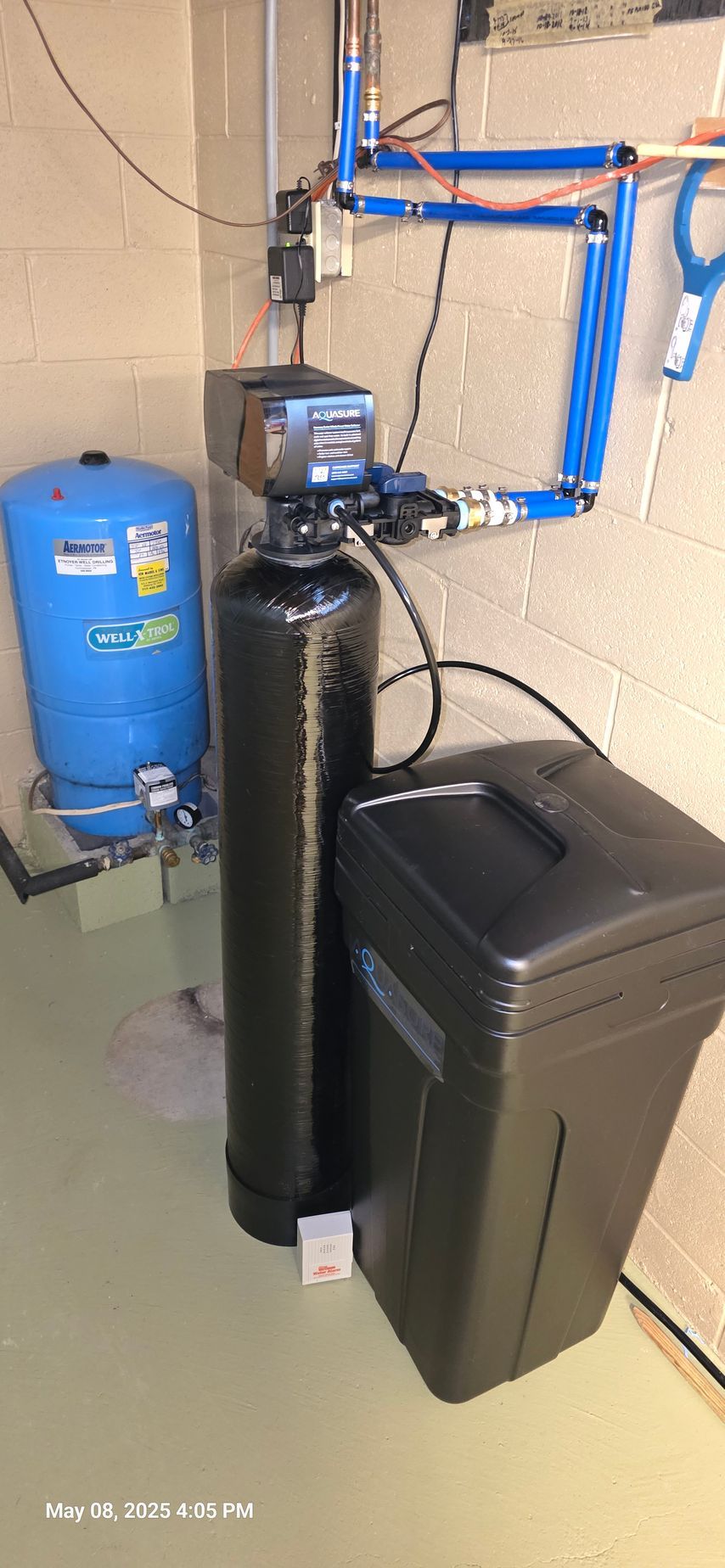 Water filtration system with blue pressure tank, black filter tank, and salt tank, installed in a basement.