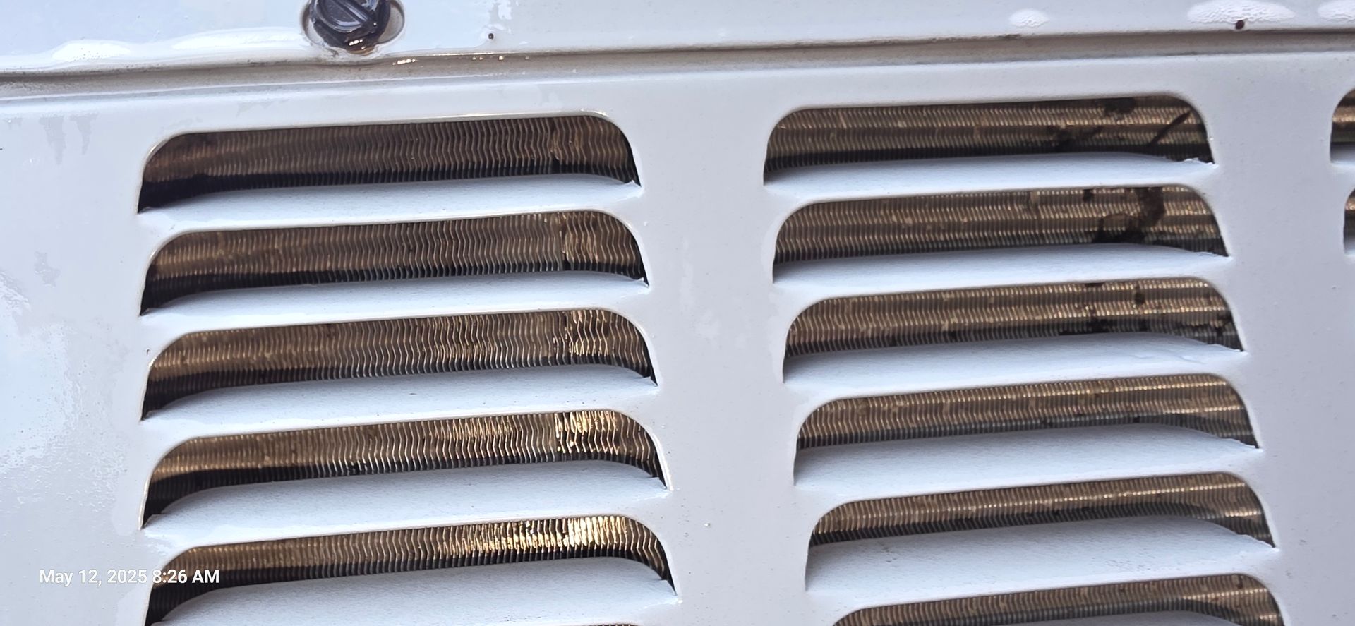 Close-up of a white vent with rectangular slots; the slots are filled with brown grime. A small, black dial sits above the vent.