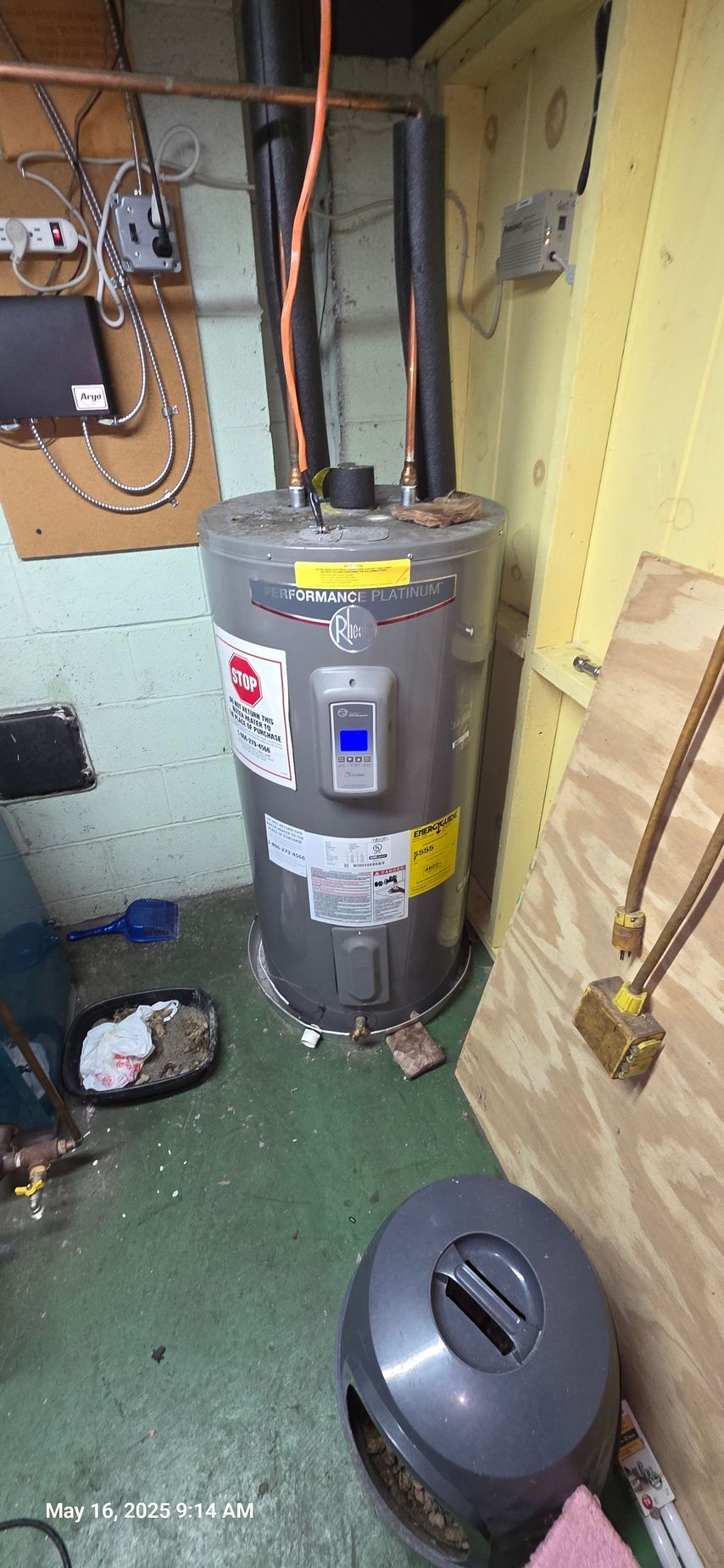 Water heater in a basement. The unit is silver and tall, with black pipes leading up. The room has a green floor.