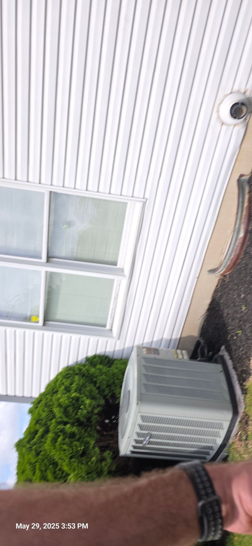 An air conditioning unit next to a green bush, next to a white house with windows.