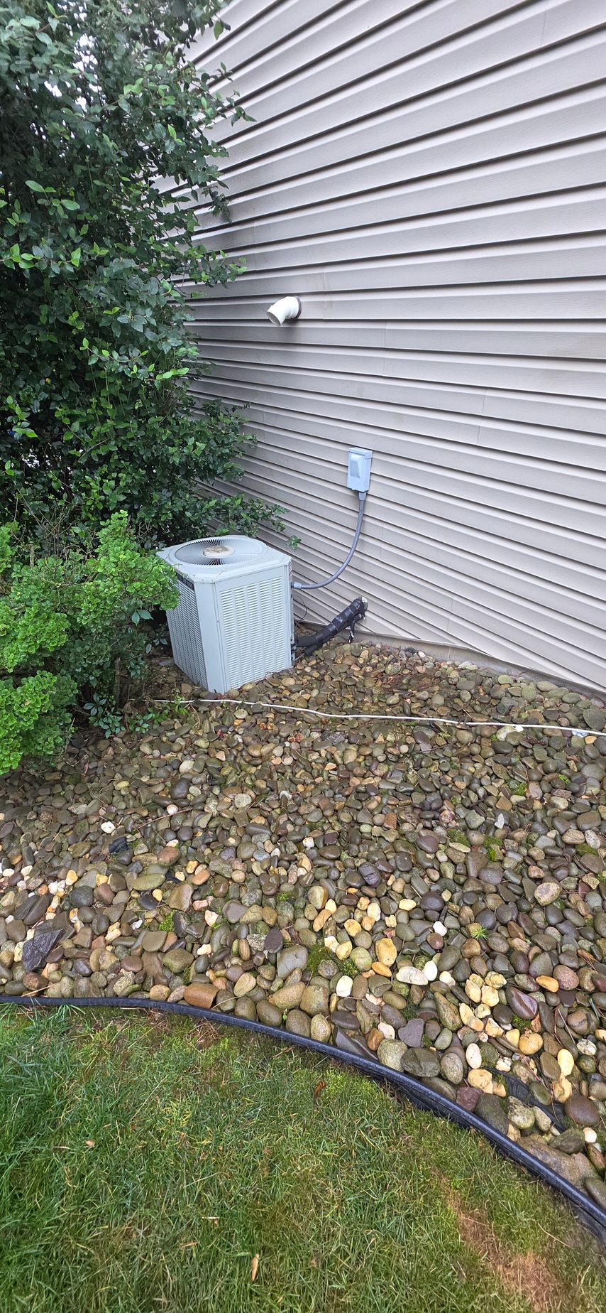 An air conditioning unit next to a house with gravel, shrubs, and grass.