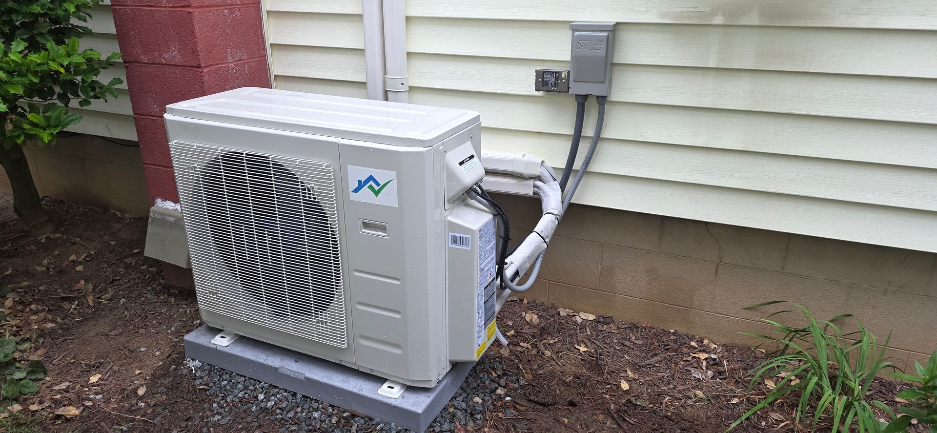 An outdoor heat pump unit next to a house. The unit is beige, with a metal grate and a logo.