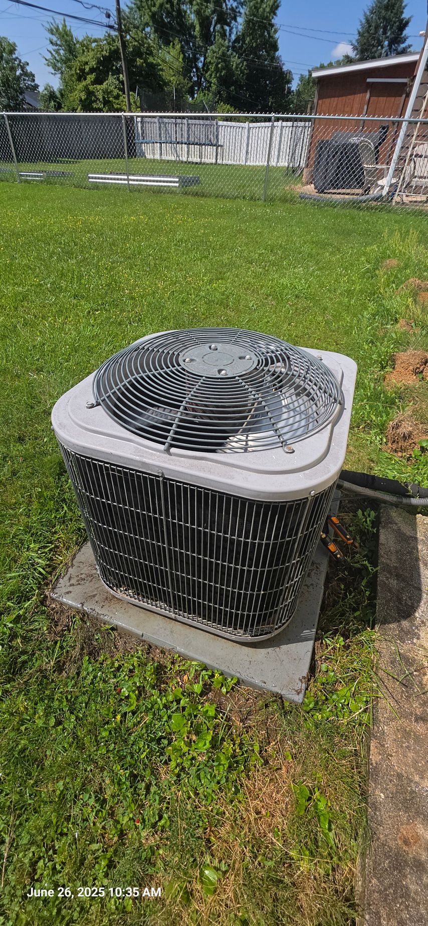 Air conditioner unit in a backyard, surrounded by grass and a fence.