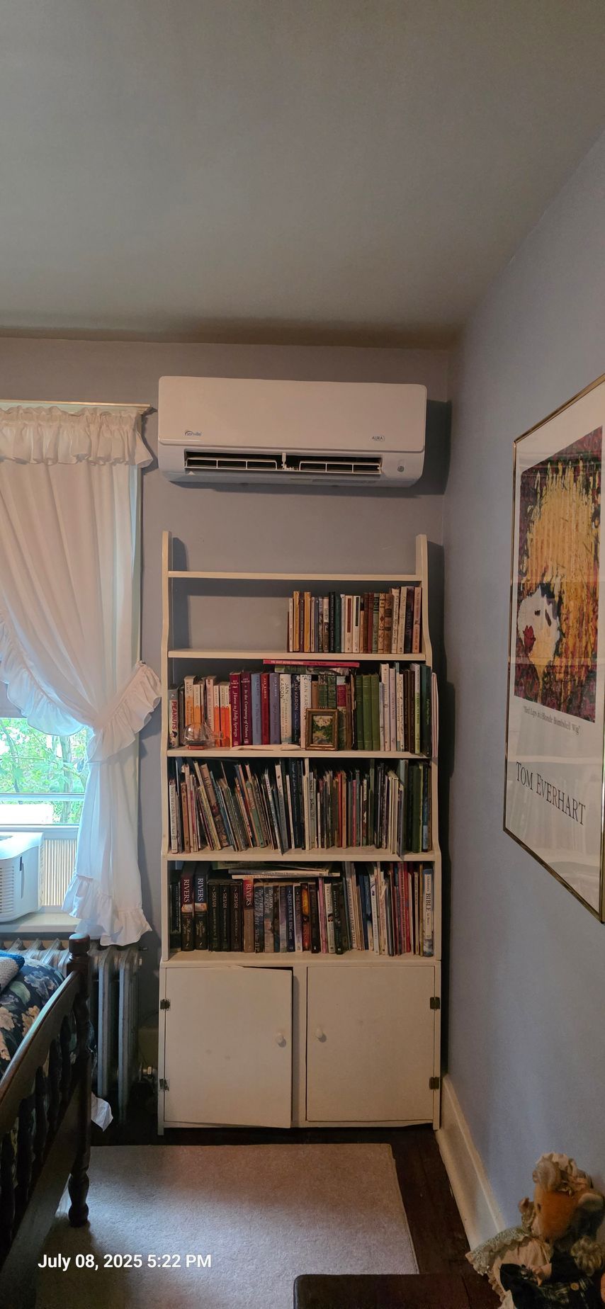 White bookshelf filled with books, an air conditioner above. A poster on the wall.
