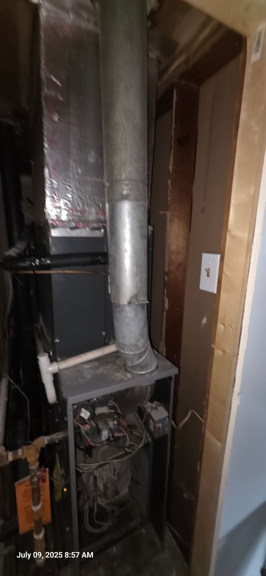 Furnace with metal vent in an enclosed area with light-colored walls and electrical outlet.