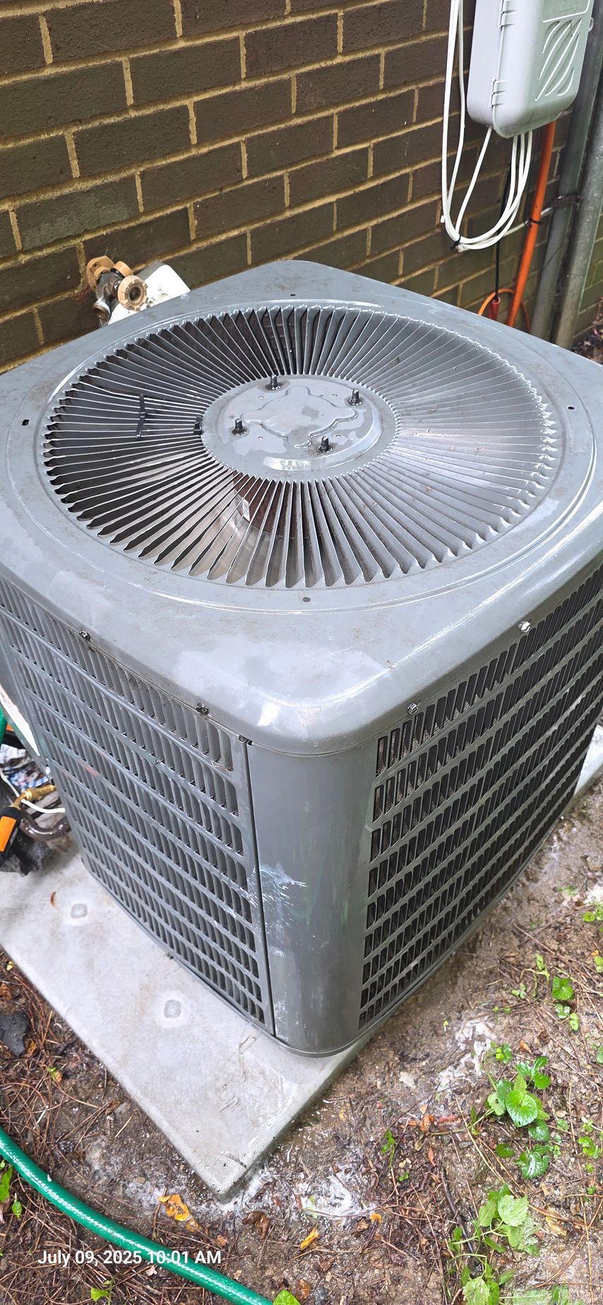 An air conditioning unit outdoors, sitting on a concrete pad next to a brick wall.