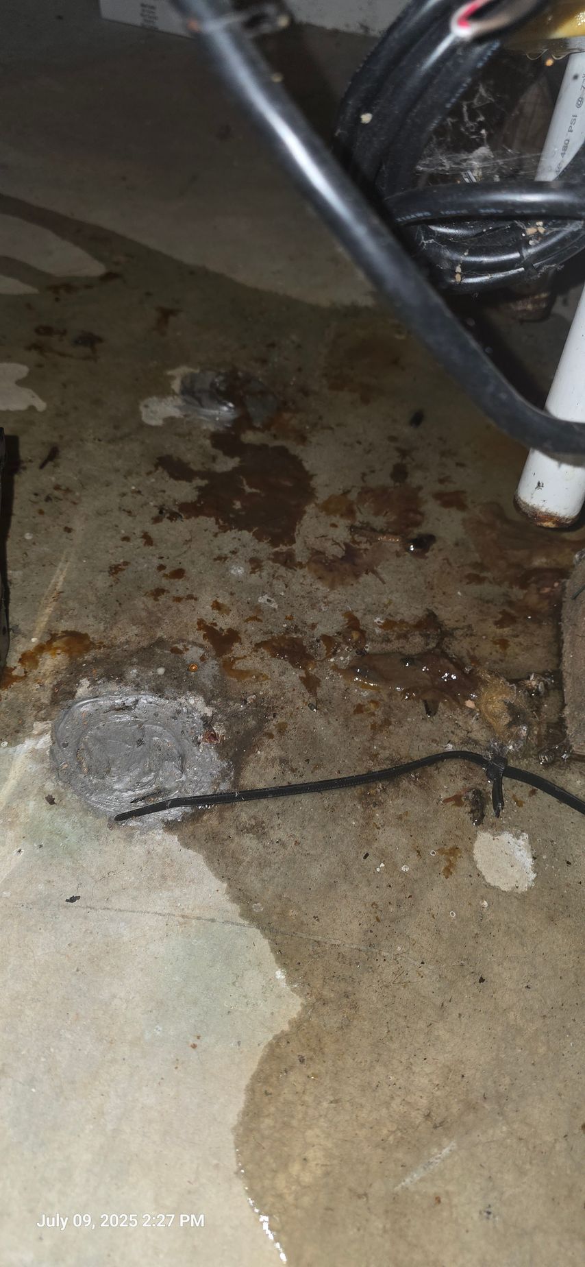 Concrete floor with rust-colored stains and wet spots, black cables and white pipe.
