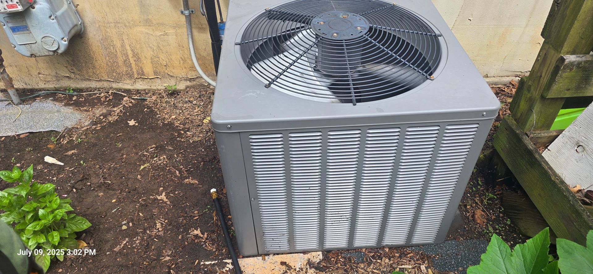 An air conditioning unit sits outdoors near a wall and a wooden structure, surrounded by dirt and plants.