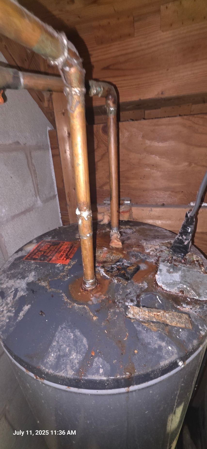 A corroded water heater with copper pipes in a cramped space.