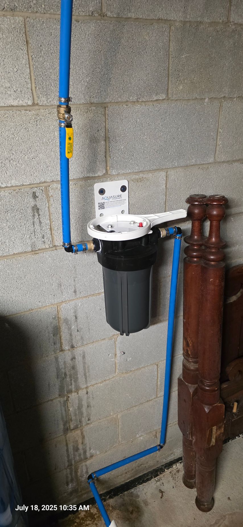 Blue plumbing pipes connected to a water filter attached to a cinder block wall, with a bed frame in the background.