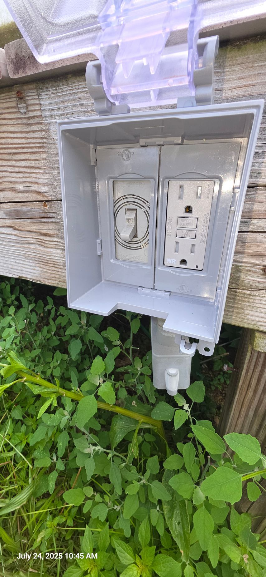 Outdoor electrical outlet box, gray with open clear plastic cover, mounted on wood, with green vegetation in the foreground.